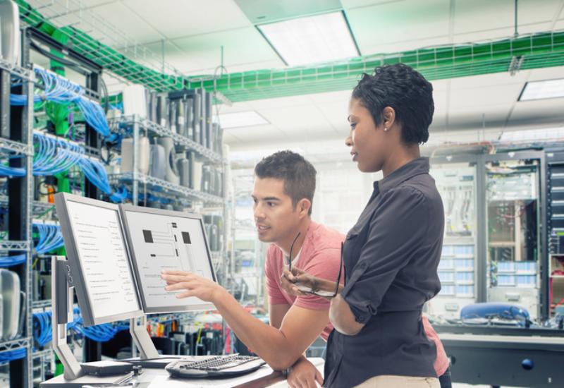 A man and a woman looking at a pair of computer monitors in a room with computer wires