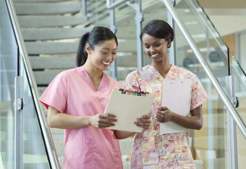 Two female nurses wearing pinks scrubs looking at a medical chart together and smiling