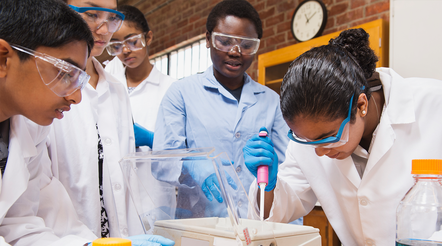 A group of five students working at a lab table wearing lab coats and safety goggles